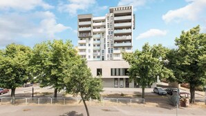 Un immeuble résidentiel à plusieurs étages, doté de balcons, d’un espace de stationnement et d’arbres au premier plan.