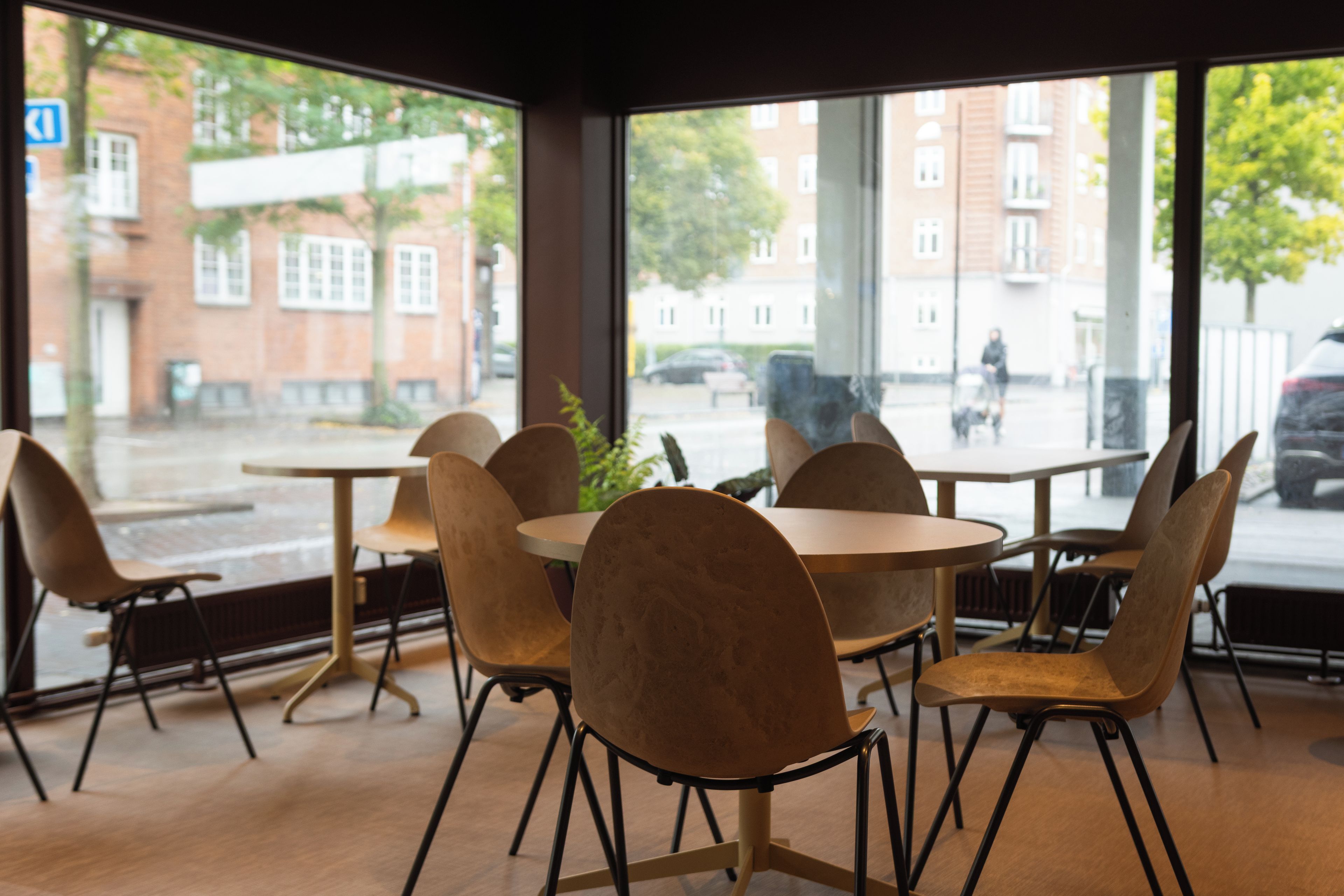 Un espace de détente intérieur avec des tables et des chaises rondes, de grandes fenêtres donnant sur une rue et des immeubles.