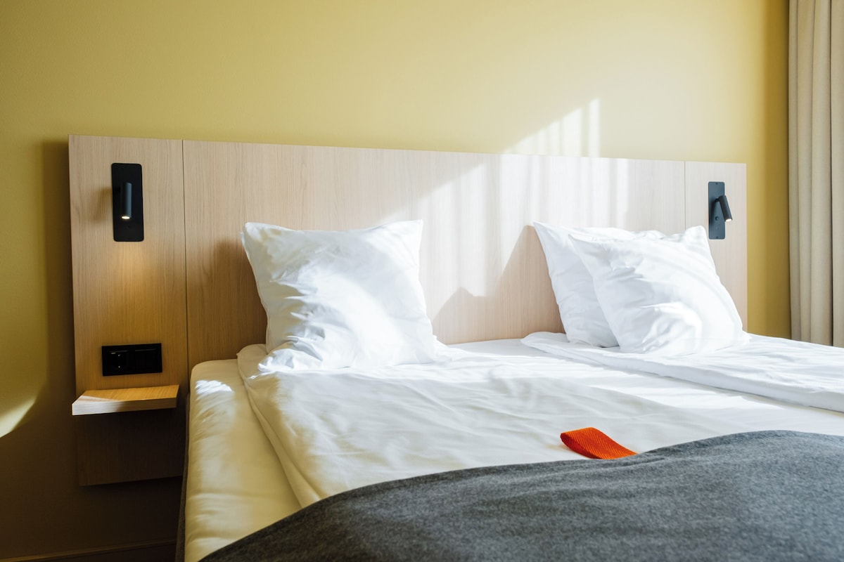 A hotel room with a wooden headboard, white bedding, and a bedside table with a lamp.