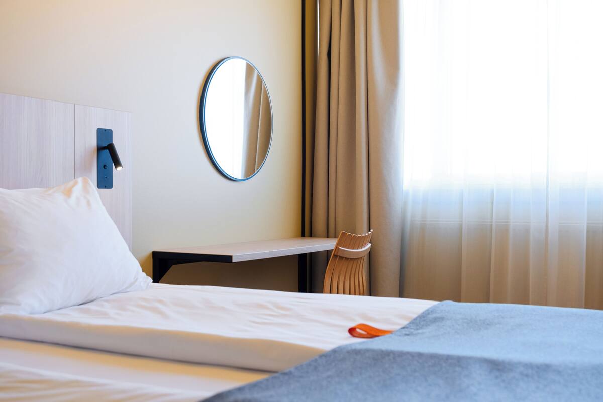 Quarto de hotel com cama, mesa de cabeceira, espelho e janela com cortinas.