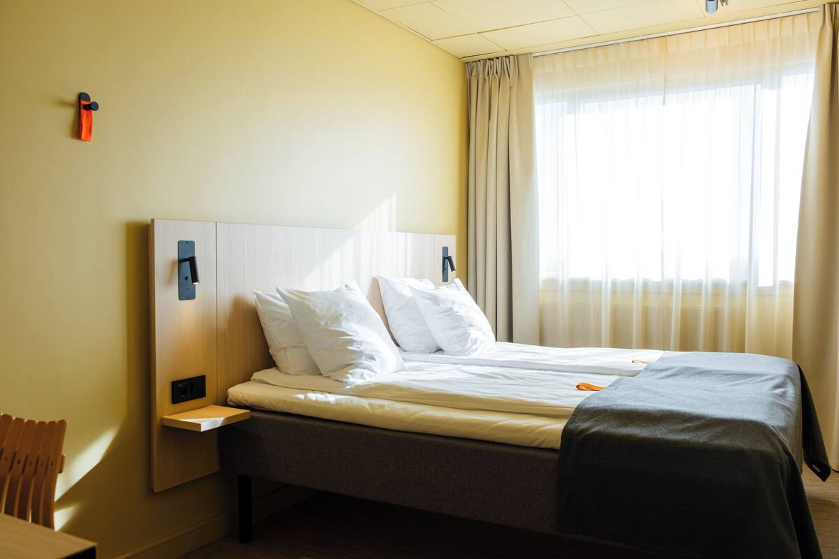 Quarto de hotel com uma cama, mesa de cabeceira, abajur, janela com cortinas e um porta-toalhas fixado na parede.