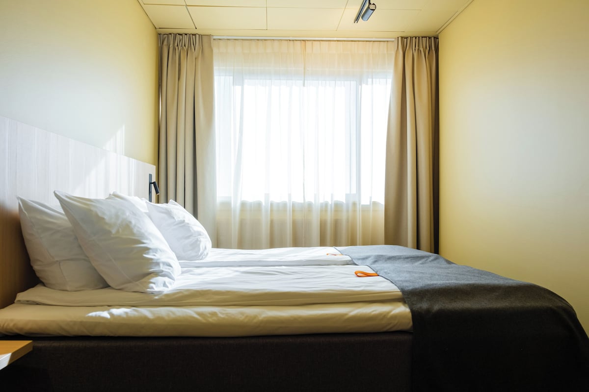 A hotel room with a bed, pillows, a blanket, a pair of scissors, and a window with curtains.