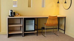 A compact hotel room with a desk, a chair, a mini-fridge, a coffee machine, and a wall-mounted clock.