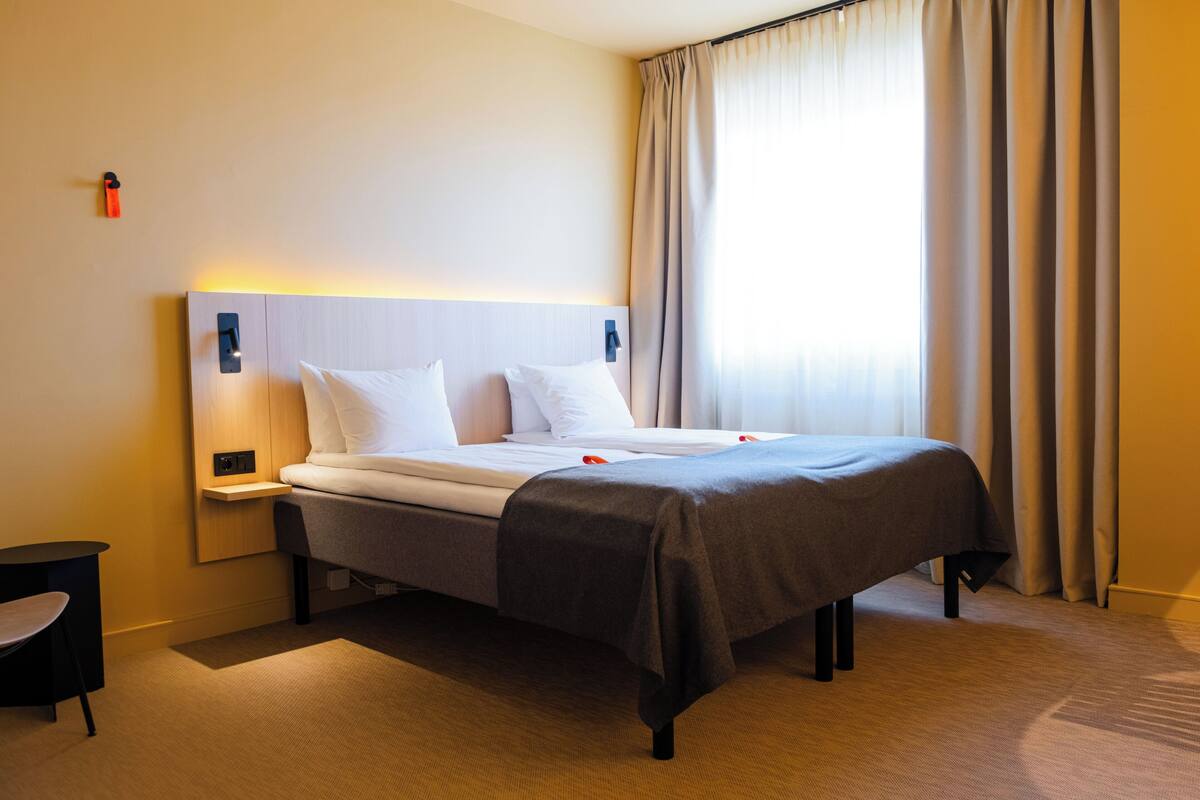 Quarto de hotel com cama, mesas de cabeceira, uma poltrona e janela com cortinas.