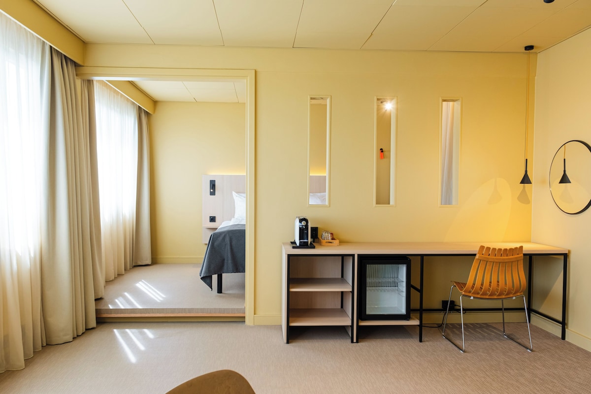 A hotel room with a bed, a desk, a chair, and a small refrigerator.