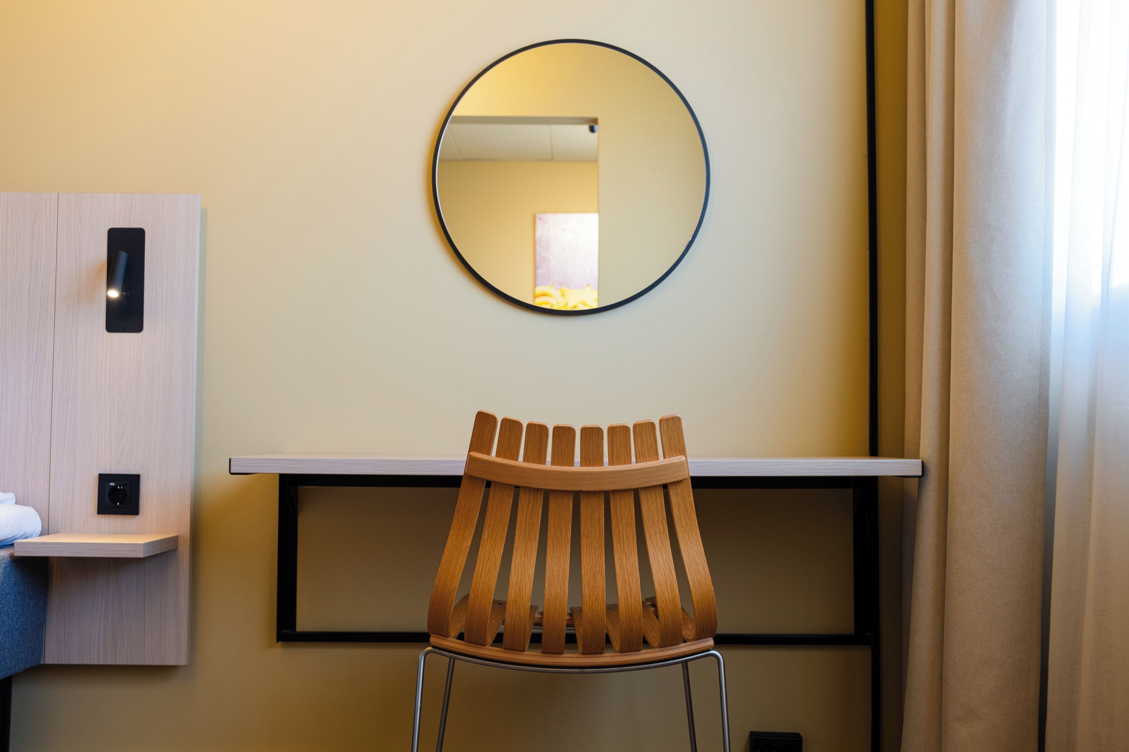 Habitación de hotel con una silla de madera, un espejo redondo en la pared y un pequeño estante con una toalla.
