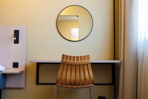 Hotellihuone, jossa on puinen tuoli, pyöreä peili seinällä ja pieni hylly, jonka päällä on pyyhe.