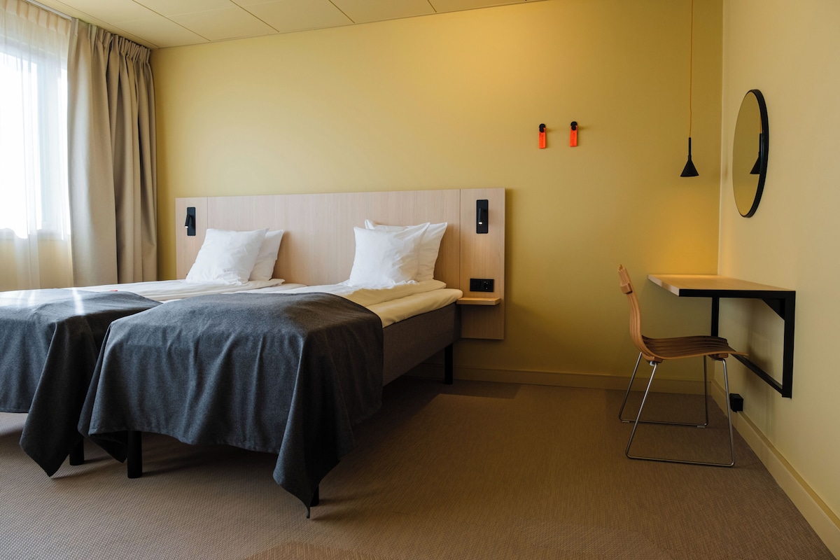 A hotel room with two beds, a desk, a chair, a mirror, and a wall-mounted light fixture.