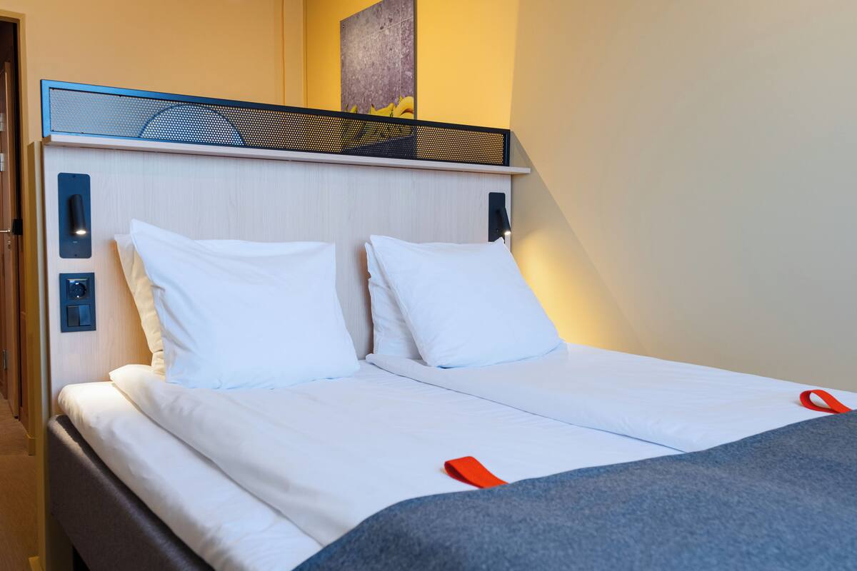 Uma cama de casal com roupa de cama branca, cabeceira azul e um objeto vermelho na cama.