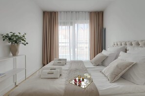 Ein Hotelzimmer mit Bett, Nachttisch, Blumentopf, einem Fenster mit Vorhängen und einem Tablett mit Früchten und Getränken.