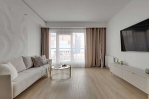 Ein modernes Wohnzimmer mit einer Couch, einem runden Couchtisch, einem Fernseher und Blick durch große Fenster.
