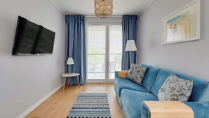 A modern living room with a blue sofa, a flat-screen TV, a wicker ceiling light, and a framed picture on the wall. - Navio with Balcony | Seaside (Gdańsk)