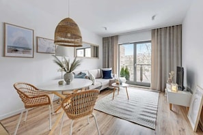 A modern living room with a round dining table, wicker chairs, a sofa, a TV, and large windows with curtains. - Waterlane F13 | Spa, Pool & Gym | Riverside (Gdańsk)