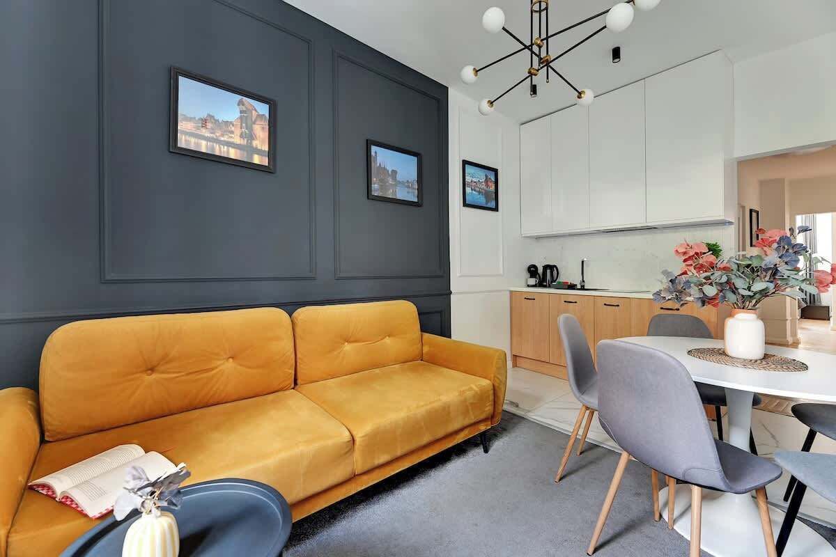 A modern living room with a sofa, dining table, chairs, and a kitchen area with cabinets and a vase of flowers.