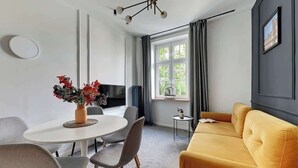 Ein modernes Wohnzimmer mit einem runden Esstisch, grauen Stühlen, einem gelben Sofa, einem Fernseher und einem Fenster mit Vorhängen.