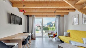 En moderne stue med træloft, en gul sofa, et spisebord med stole og et TV ophængt på væggen.