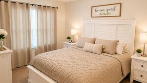 Un dormitorio con cama, mesitas de noche, una mesilla, un reloj de pared, un cartel enmarcado y una ventana con cortinas.