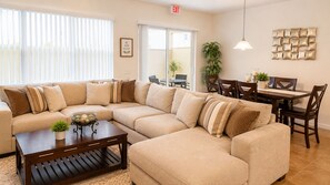 A living room with a sectional sofa, a coffee table, a dining table with chairs, and a wall-mounted light fixture. - Townhome Close to Disney w Resort Pool (Kissimmee)