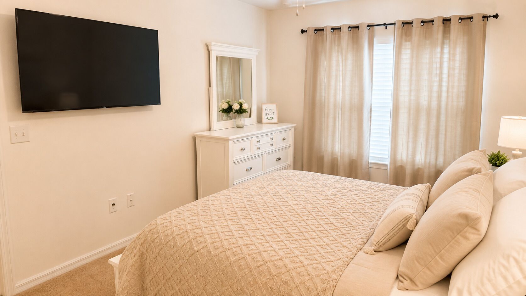 A bedroom with a bed, a dresser, a mirror, a flat-screen TV, and a window with curtains.