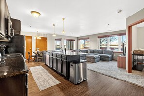 A modern kitchen with stainless steel appliances, a dining area with a table and chairs, and a living room with a gray sectional sofa.