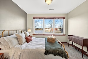 Une chambre à coucher avec un lit, une table de chevet, une lampe, une fenêtre donnant sur un beau paysage et un banc.