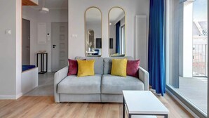 A modern living room with a grey sofa, two mirrors, a white table, and a view through a large window. - Grano Residence 64 with Balcony | Riverside (Gdańsk)