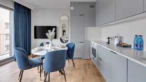 A modern kitchen with grey cabinets, a dining area with blue chairs, a flat-screen TV, and a large window with blue curtains. - Grano Residence 64 with Balcony | Riverside (Gdańsk)
