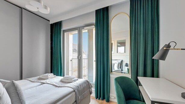 A hotel room with a bed, a desk, a chair, a mirror, and a view through a glass door. - Grano Residence 64 with Balcony | Riverside (Gdańsk)