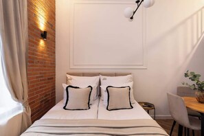 Quarto com parede de tijolos, cama com roupa de cama branca e almofadas com detalhes pretos, mesa de cabeceira, cadeira e luminária pendente.