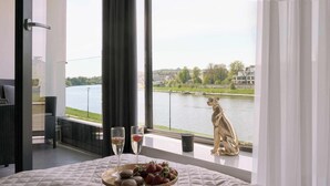 A bedroom with a bed, a tray of strawberries, two glasses of champagne, and a statue by the window overlooking a river. - Vistula View - Riva Verona | Parking (Kraków)