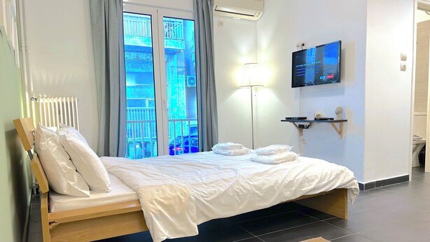 A hotel room with a bed, a TV, a window with a view of the street, and a radiator. - Cozy Studio Near City Center I Walk to Metro (Athina)