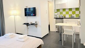 A hotel room with a bed, a desk with a chair, a television mounted on the wall, a small table with a lamp, and a kitchenette area. - Cozy Studio Near City Center I Walk to Metro (Athina)