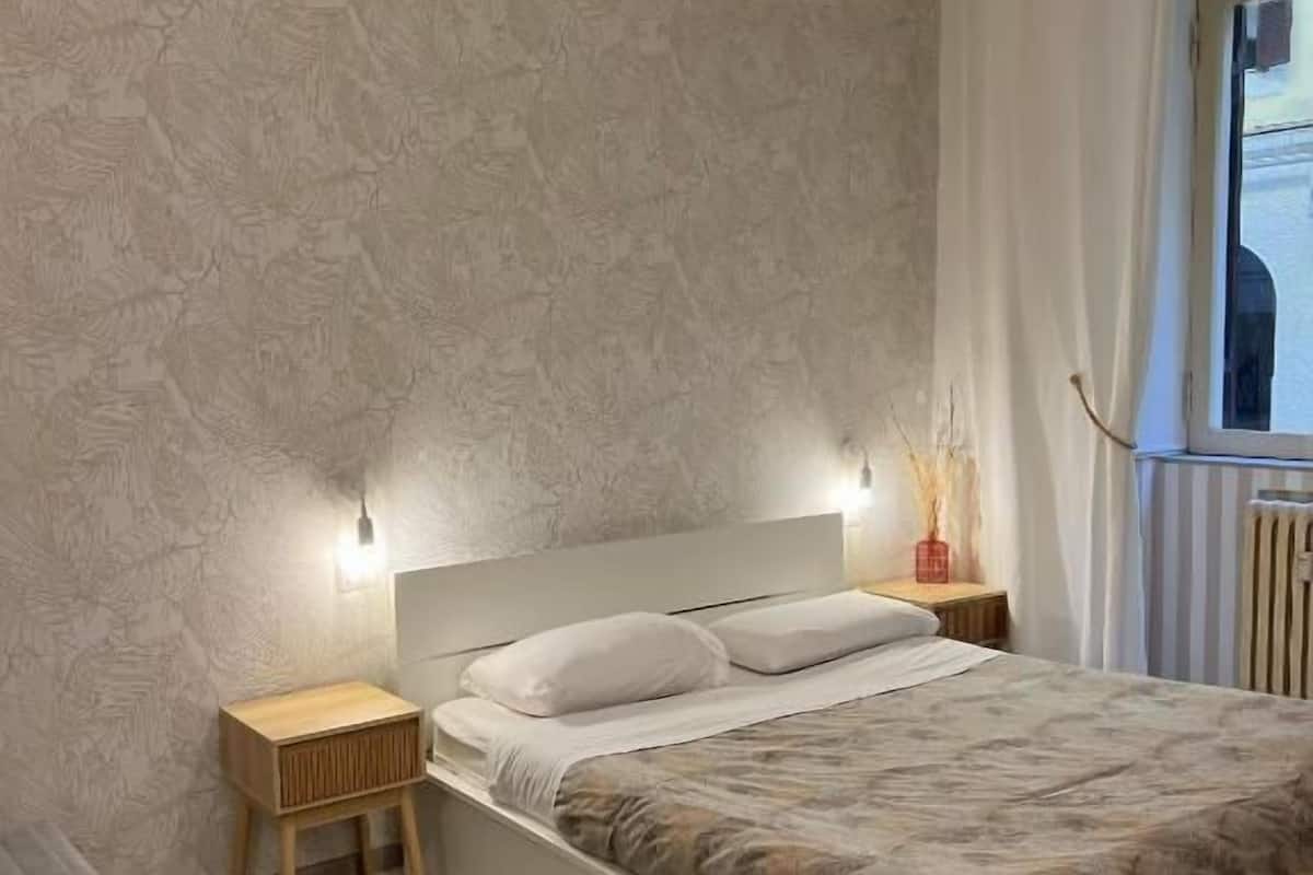 Un dormitorio con cama, mesitas de noche, lámpara colgante, ventana con cortinas y un radiador.