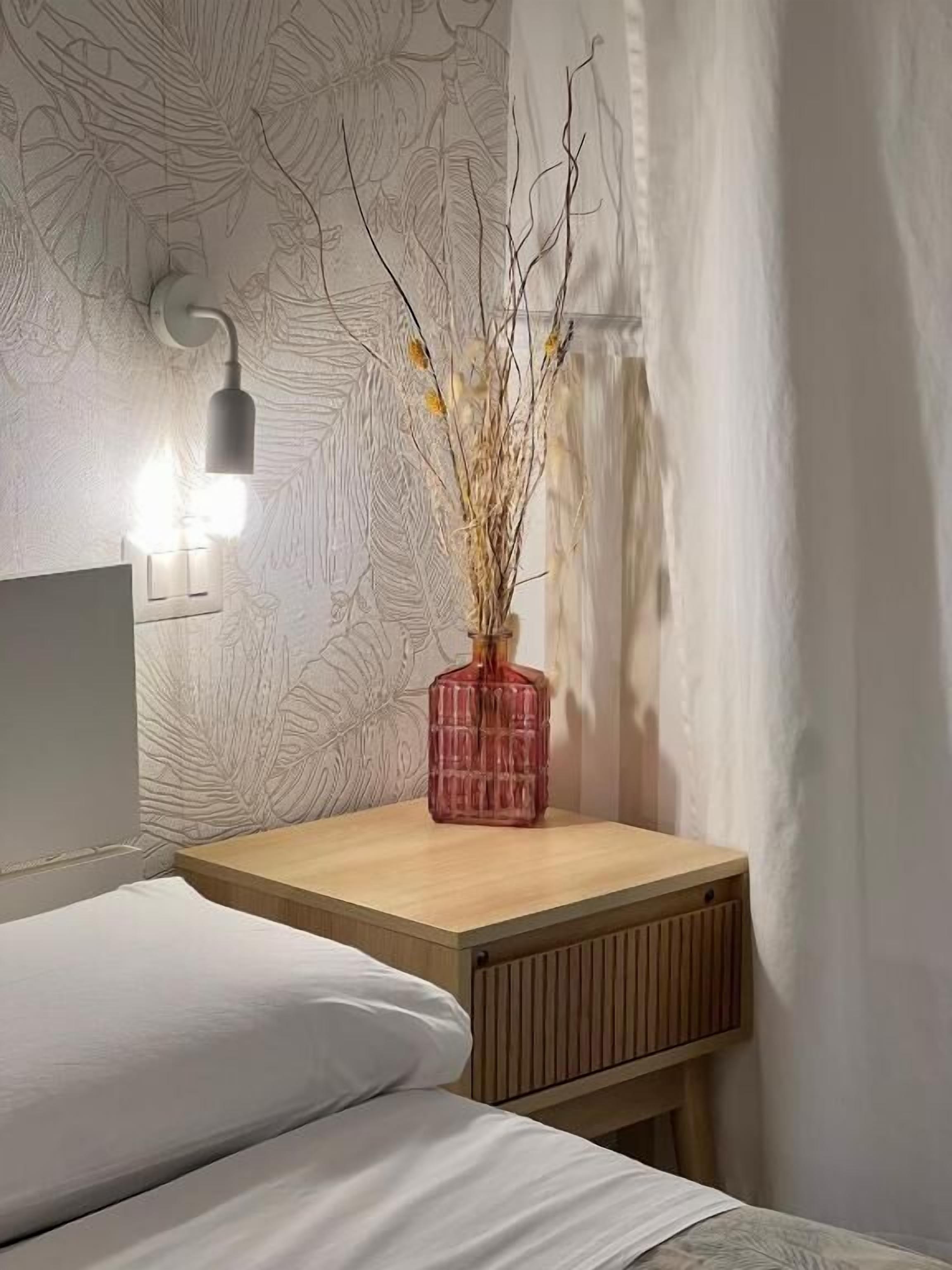 A bedroom with a bed, a bedside table with a lamp, and a vase with branches.