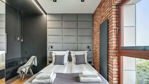 A modern hotel room with a large bed, a brick accent wall, a large window, and a shower area. - Nowa Motława 32.1 | River View | SPA & Parking (Gdańsk)