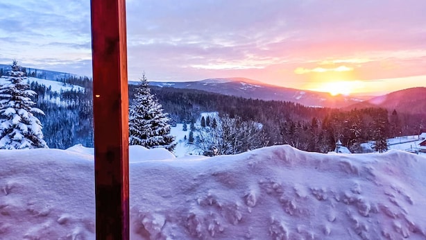 Schneebedeckte Landschaft mit Sonnenuntergang, sichtbaren Bergen und einer Holzhütte.