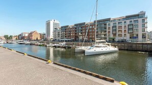 Ein Yachthafen mit vertäuten Booten, eine Uferpromenade und moderne Gebäude im Hintergrund.