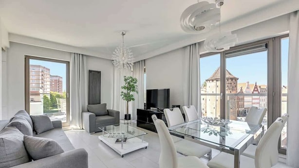 Ein modernes Wohnzimmer mit Glastisch, grauem Sofa und einem Essbereich mit weißen Stühlen.