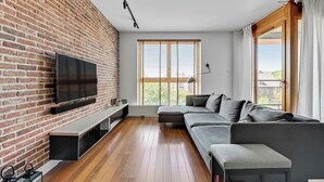 A modern living room with a brick wall, a flat-screen TV, a gray sofa, a wooden floor, and a large window with blinds. - Aura 29 | Riverside | City Center (Gdańsk)