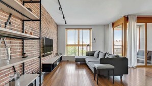 A modern living room with a brick wall, wooden flooring, a sectional sofa, a TV, and a large window. - Aura 29 | Riverside | City Center (Gdańsk)