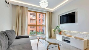 A modern living room with a sofa, a glass coffee table, a TV mounted on the wall, and a window with curtains. - Aura 99B | Riverside | City Center (Gdańsk)
