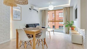 A modern living room with a dining table, chairs, a sofa, a potted plant, and a large window with curtains. - Aura 99B | Riverside | City Center (Gdańsk)