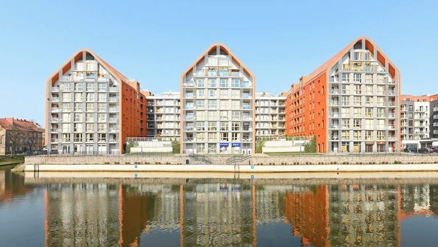 A modern hotel with a unique triangular roof design, located beside a calm body of water. - Aura 99B | Riverside | City Center (Gdańsk)