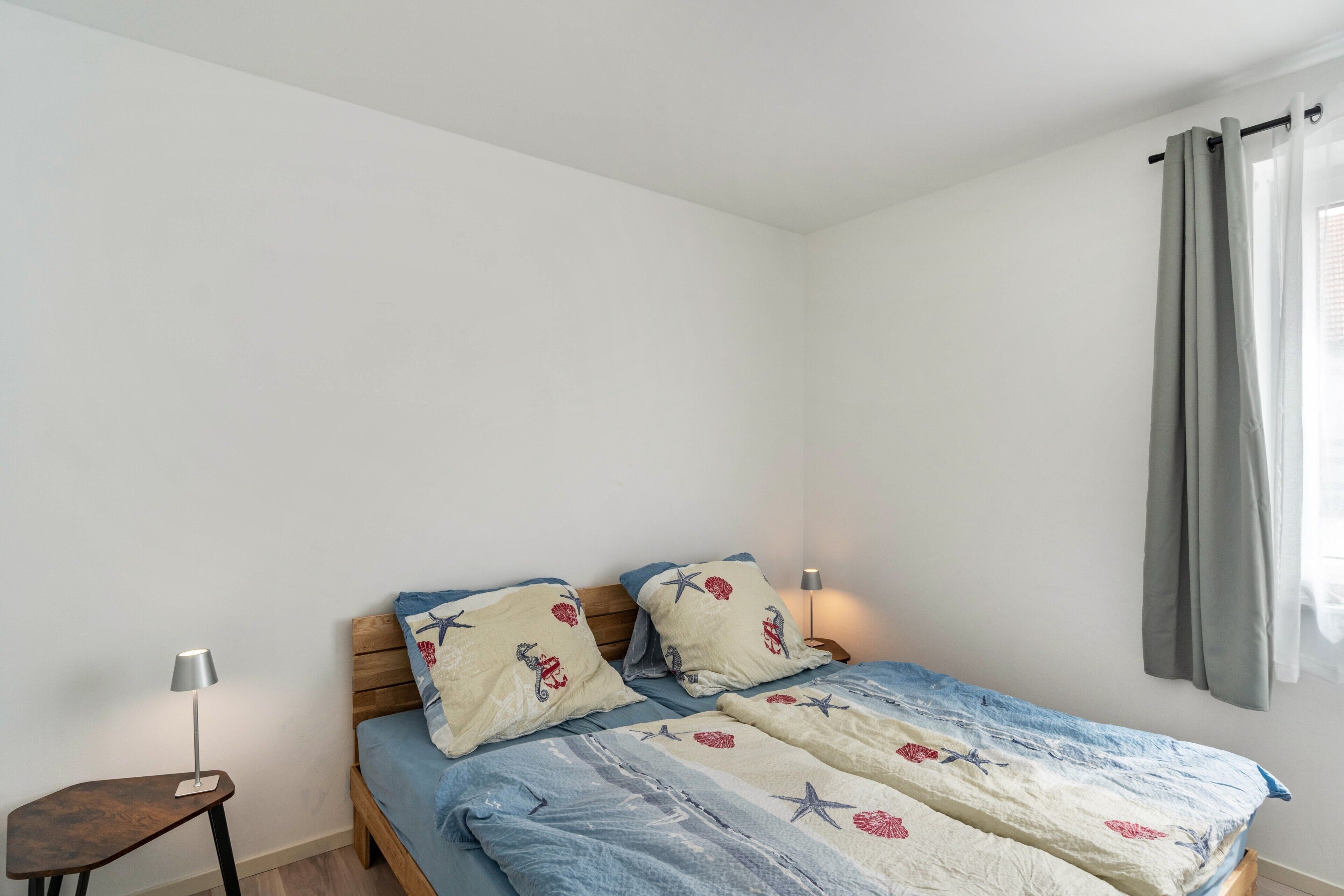 Ein Einzelbett mit blau-weiß gestreifter Bettdecke, zwei Kissen mit Meeresmotiven, ein Nachttisch mit Lampe und ein Fenster mit grauen Vorhängen.