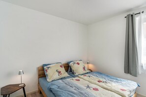 A single bed with a blue and white striped bedspread, two pillows with a nautical theme, a bedside table with a lamp, and a window with grey curtains.