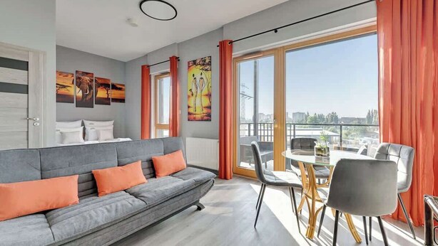 Ett modernt vardagsrum med en grå soffa, orange kuddar, ett matbord med stolar och stora fönster med orange gardiner.