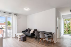 Una sala de estar moderna con un sofá, una mesa de comedor y sillas. Hay una pequeña mesa blanca y una mesita de noche con una lámpara. La habitación tiene ventanas grandes con cortinas y vistas al exterior.