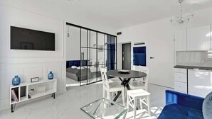 A modern living room with a glass coffee table, white furniture, a blue sofa, and a kitchen area with white cabinets. - Długa Street | Main Old Town (Gdańsk)