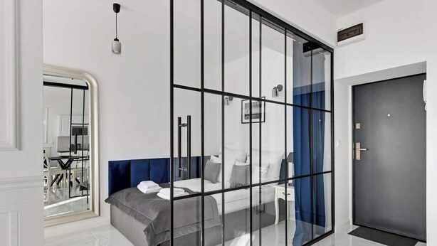 A modern hotel room with a glass partition, a bed, a bedside table, a wardrobe, and a door. - Długa Street | Main Old Town (Gdańsk)