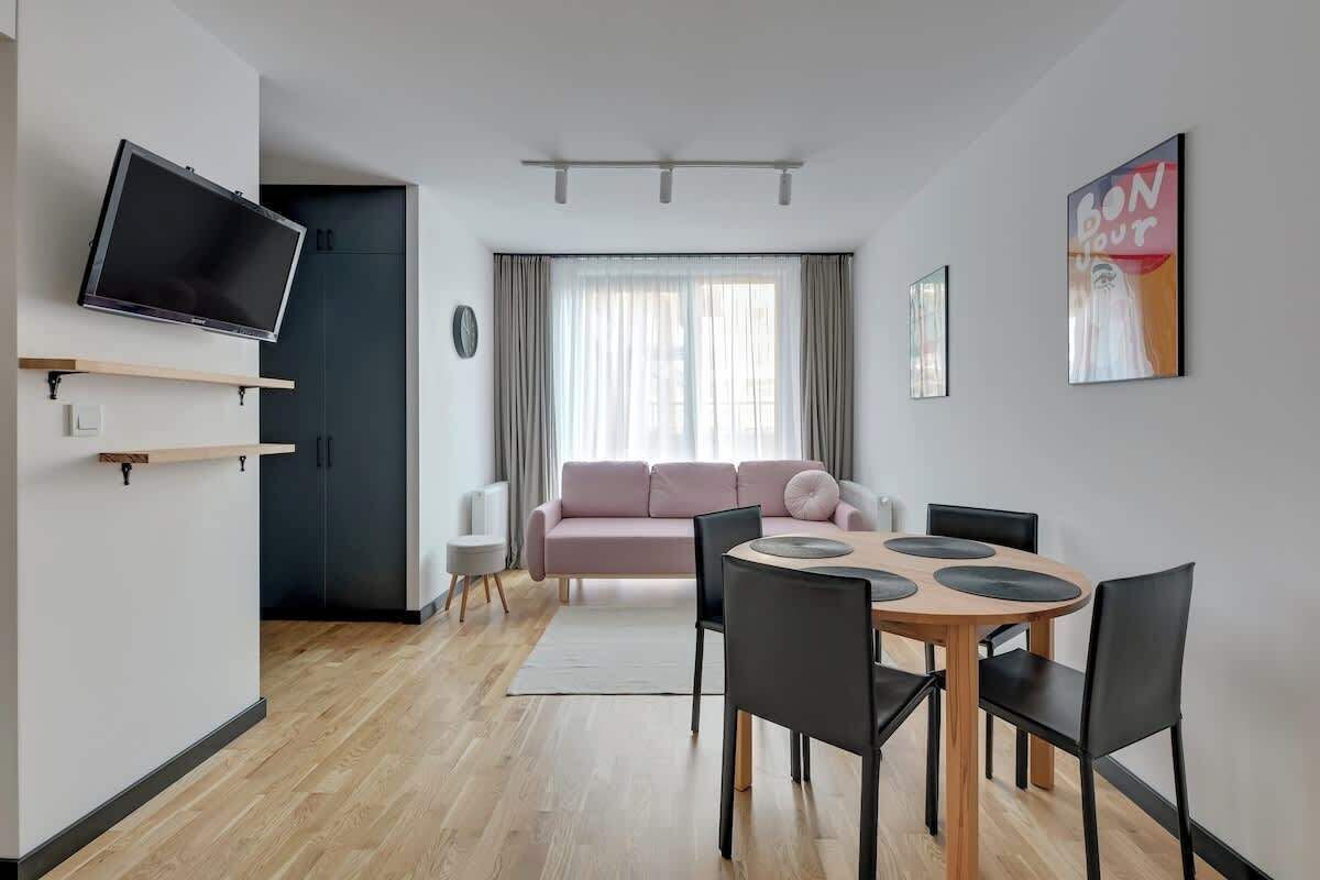 Ein modernes Wohnzimmer mit einem Flachbildfernseher, einem rosafarbenen Sofa, einem Esstisch mit Stühlen und einem großen Fenster mit Vorhängen.